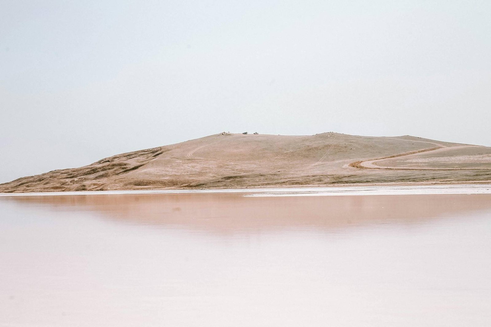 A gentle, barren hill rises behind a calm, pale pink lake under a cloudy sky. A winding dirt path curves up the hill, and the landscape appears serene and minimalistic.